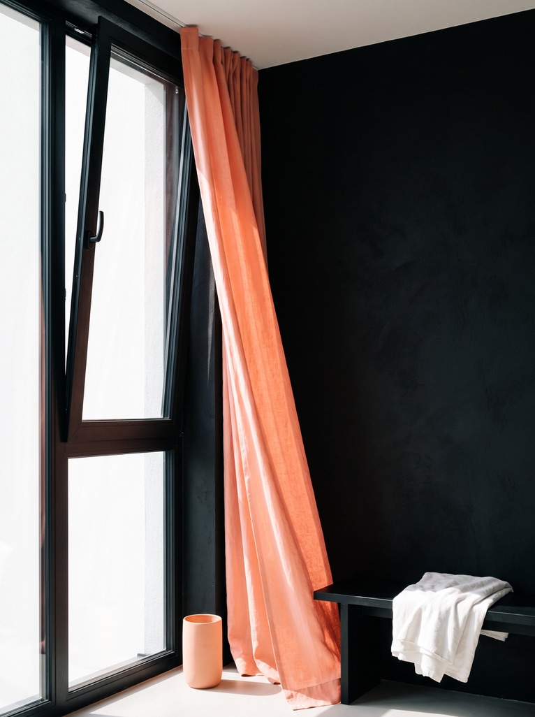 Tall window with a peach curtain blowing inward beside a black bench